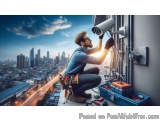 The image shows a man working on a security camera mounted on the side of a building. He is wearing a blue shirt, jeans, and...