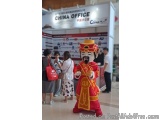 The image shows a person dressed in a traditional Chinese costume, standing in front of a banner that reads "China Office". T...