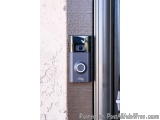 The image shows a Ring video doorbell mounted on the side of a door. The doorbell is black in color and has a round button in...