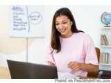 The image shows a young woman sitting at a desk with a laptop in front of her. She is wearing a pink t-shirt and has long bro...