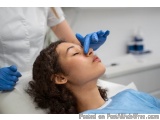 The image shows a young woman lying on a white surface with her eyes closed, receiving a facial treatment. She is wearing a b...