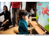 The image shows a young child, probably around 2-3 years old, standing in front of a large whiteboard with a colorful illustr...