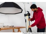 The image shows a man in a red hoodie standing in front of a white wall with a black umbrella on the left side. He is holding...