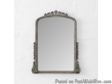 The image is of an ornate, rectangular mirror hanging on a white brick wall. The mirror is made of silver metal and has an in...