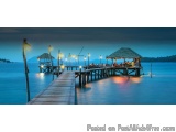 The image shows a long wooden pier extending into the ocean at night. The pier is made of wood and has a thatched roof hut on...