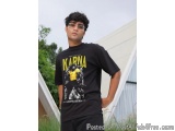 The image shows a young man standing in front of a white building with greenery in the background. He is wearing a black t-sh...