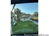 The image shows a view from the driver's seat of a vehicle, looking out the window. Through the window, we can see a house th...