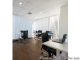 The image shows an empty office space with multiple desks and chairs arranged in a row. The desks are white and have laptops...