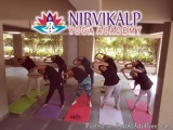 The image shows a group of people performing a yoga class at Nirvana Kalp Yoga Academy. The class is taking place in a large...