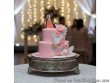 The image is of a three-tiered pink cake on a silver cake stand. The cake is decorated with white fondant flowers and silver...