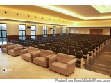 The image shows a large auditorium with rows of beige leather sofas arranged in neat rows. The room has a high ceiling with r...