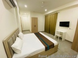 The image shows a modern and spacious bedroom with a large bed in the center. The bed has a beige headboard and white linens...