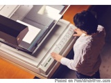 The image shows a person standing in front of a large printing machine. The person is wearing a white shirt and appears to be...