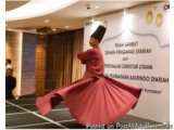 The image shows a young man performing a traditional dance in a large room with a banner in the background. The man is wearin...