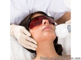 The image shows a young woman lying down on a white surface with her eyes closed, receiving a laser treatment on her face. Sh...
