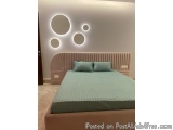 The image shows a modern bedroom with a large bed in the center. The bed has a light pink headboard with a curved design and...