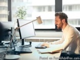 The image shows a man sitting at a desk in front of a computer monitor. He is wearing a beige shirt and has a beard and is sm...