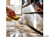 The image shows a person's hand holding a yellow sponge and cleaning a kitchen countertop. The countertop appears to be dirty...