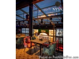 The image shows the interior of a restaurant named NYSA. The restaurant has a high ceiling with a skylight that allows natura...
