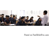 The image shows a classroom with a group of students sitting at desks and listening attentively to a teacher who is standing...