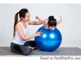 The image shows a mother and daughter playing with a blue exercise ball. The mother is sitting on a gray yoga mat and the dau...
