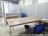 The image shows an office space with a long wooden desk in the center. The desk is surrounded by two blue office chairs on ei...
