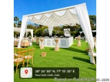 The image shows an outdoor event space with a large white canopy tent set up on a grassy lawn. The tent is decorated with whi...
