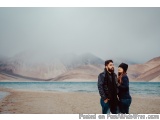The image shows a young couple standing on a sandy beach with a beautiful view of the ocean and mountains in the background....