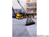 The image shows a vacuum cleaner cleaning a gray carpet in a living room. The vacuum cleaner has a long, cylindrical body wit...