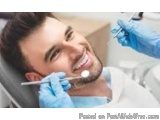 The image shows a young man sitting in a dental chair with a smile on his face. He is wearing a white shirt and blue gloves....