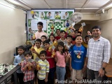 The image shows a group of children and a man posing for a photo in a classroom. The children are holding up trophies and med...