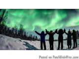 The image shows a group of six people standing on a snowy hill with their arms raised in the air, looking up at the beautiful...