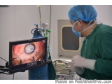 The image shows a surgeon performing an operation in an operating room. The surgeon is wearing a green surgical gown, a blue...