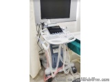 The image shows a medical equipment, specifically an ultrasound machine, in a hospital room. The machine is white in color an...