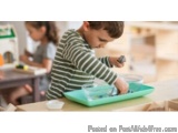 The image shows a young boy sitting at a table with a green tray in front of him. He is wearing a striped shirt and appears t...