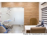 The image shows a modern dental clinic with a wooden wall on the right side and a desk on the left side. The desk has a compu...