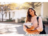 The image is a portrait of a young woman standing on a sidewalk in front of a building. She is wearing a pink long-sleeved sh...