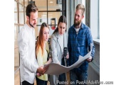 The image shows four young people, two men and two women, standing in an office or a workspace and looking at a blueprint. Th...