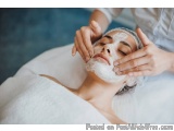 The image shows a young woman lying on a massage table with her eyes closed, receiving a facial treatment. She is covered wit...