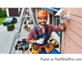 The image shows a young man wearing an orange hard hat and overalls, holding a tool belt with various tools in it. He is stan...