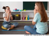 The image shows a young girl sitting on the floor in a classroom with her mother. The girl is sitting on a yellow cushion wit...