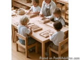 The image shows a group of four children sitting around a wooden table in a classroom. The table is covered with wooden block...