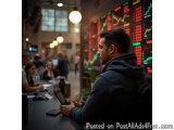 The image shows a young man sitting at a table in a trading room. He is holding a tablet in his hands and appears to be looki...