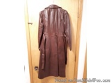 The image shows a brown leather coat hanging on a wooden door. The coat has a collar and long sleeves, and it appears to be m...