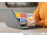 The image shows a person's hands typing on a laptop. The laptop is placed on a wooden table and the person is wearing an oran...