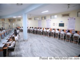 The image shows a large room with rows of desks and chairs arranged in neat rows. The desks are arranged in a semi-circle aro...