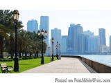 The image shows a long walkway with a row of street lamps on either side. The walkway is lined with palm trees and benches on...