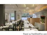 The image shows a modern and elegant dining room with a high ceiling and large windows that offer a view of the trees outside...