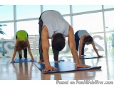 The image shows three people performing a yoga pose on blue mats in a large room with large windows. The person in the center...