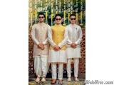 The image shows three young men standing in front of a floral archway. They are all wearing traditional Indian attire, with t...